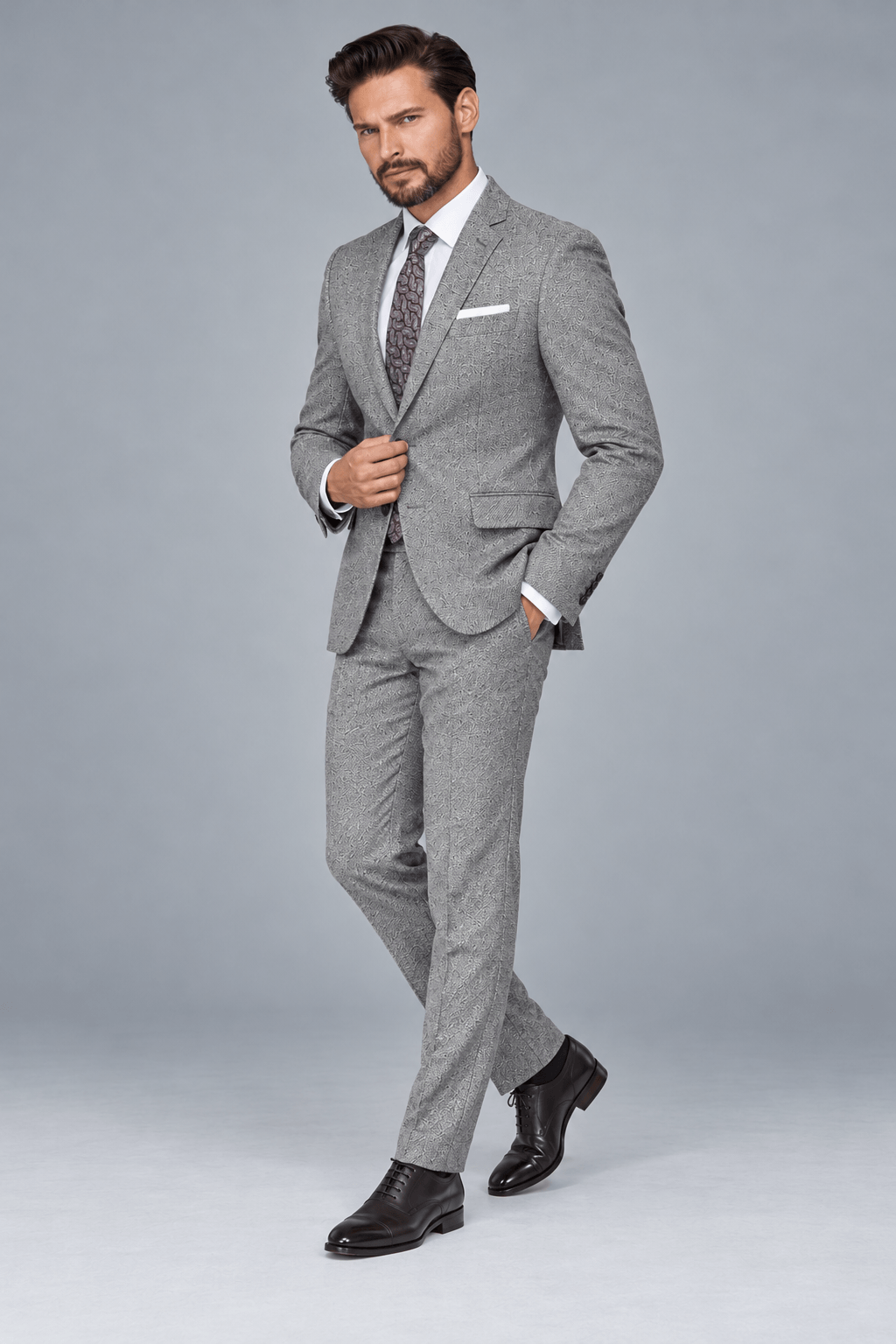 Man wearing a tailored light gray patterned two-piece suit with a white dress shirt, patterned tie, white pocket square, and black leather Oxford shoes.