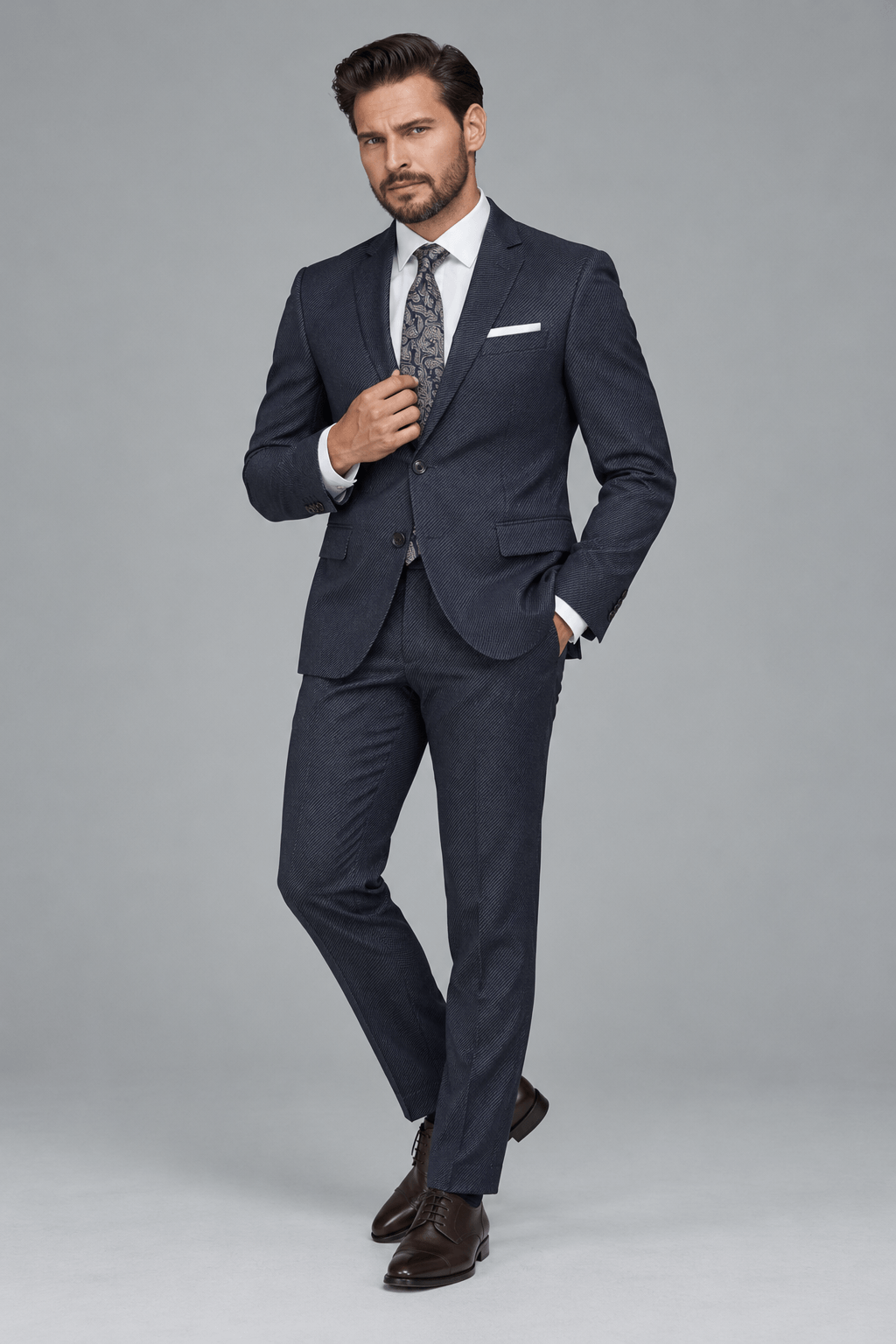 Man wearing a tailored navy blue textured two-piece suit with a white dress shirt, paisley-patterned tie, white pocket square, and dark brown leather Oxford shoes.