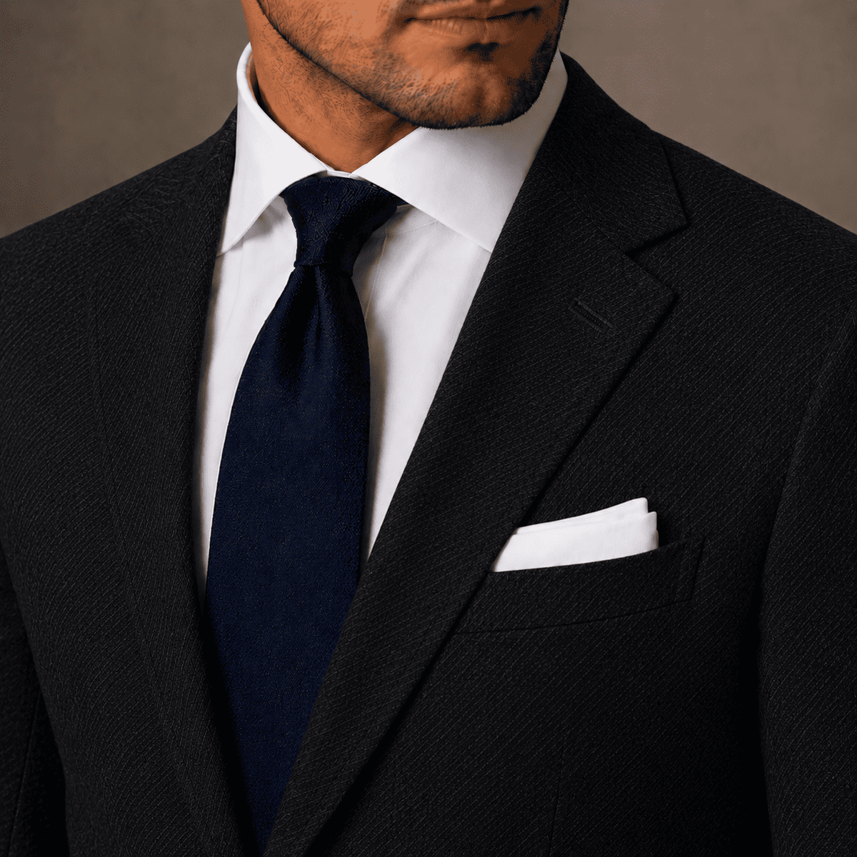Men’s black linen bespoke suit jacket with a white dress shirt, navy tie and white pocket square, highlighting the tailored lapel and textured fabric.