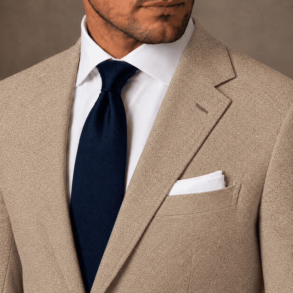 A close-up of a man wearing a beige windowpane check suit jacket, a white dress shirt, a navy blue tie, and a white pocket square.